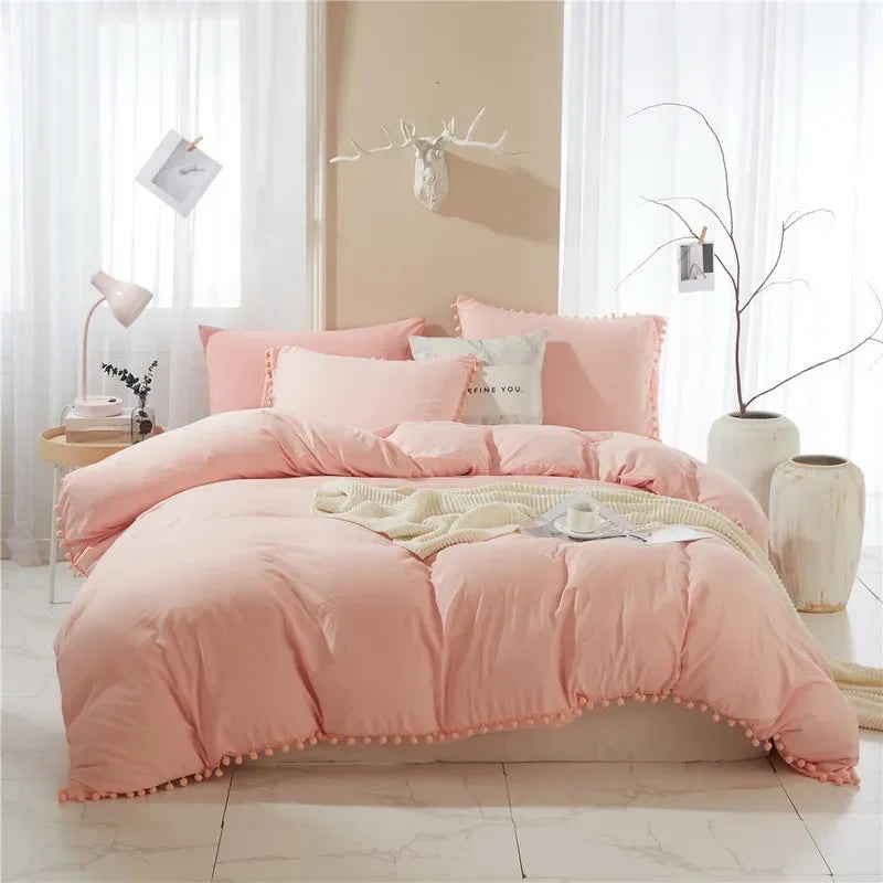 A pink duvet cover set with a pom-pom trim displayed on a bed against a light wall and white curtains.