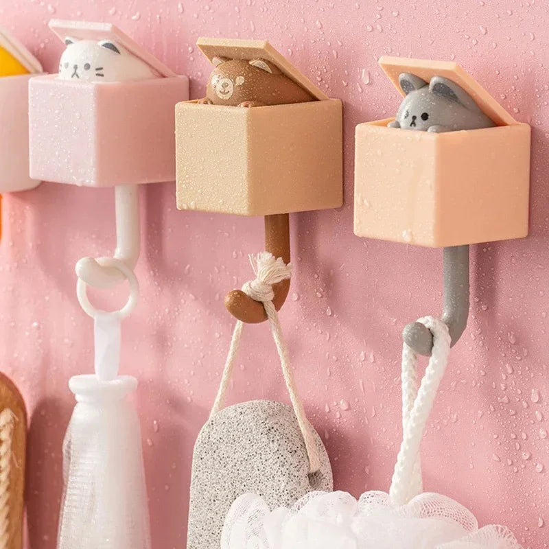 Three pink and gray cat-shaped adhesive hooks mounted on a pink wall, next to a white bottle and a gray object.