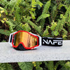 A pair of orange and black HD Motocross goggles with an anti-fog lens resting on a light surface amidst green foliage.