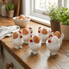 A collection of white ceramic chicken eggs with red combs and brown shells displayed on a wooden surface.