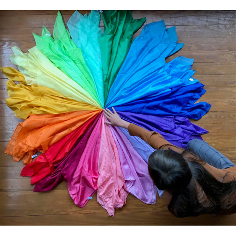 A colorful fan of Montessori Playsilks fabric in various shades of green, yellow, blue, and purple spread out on a wooden sur
