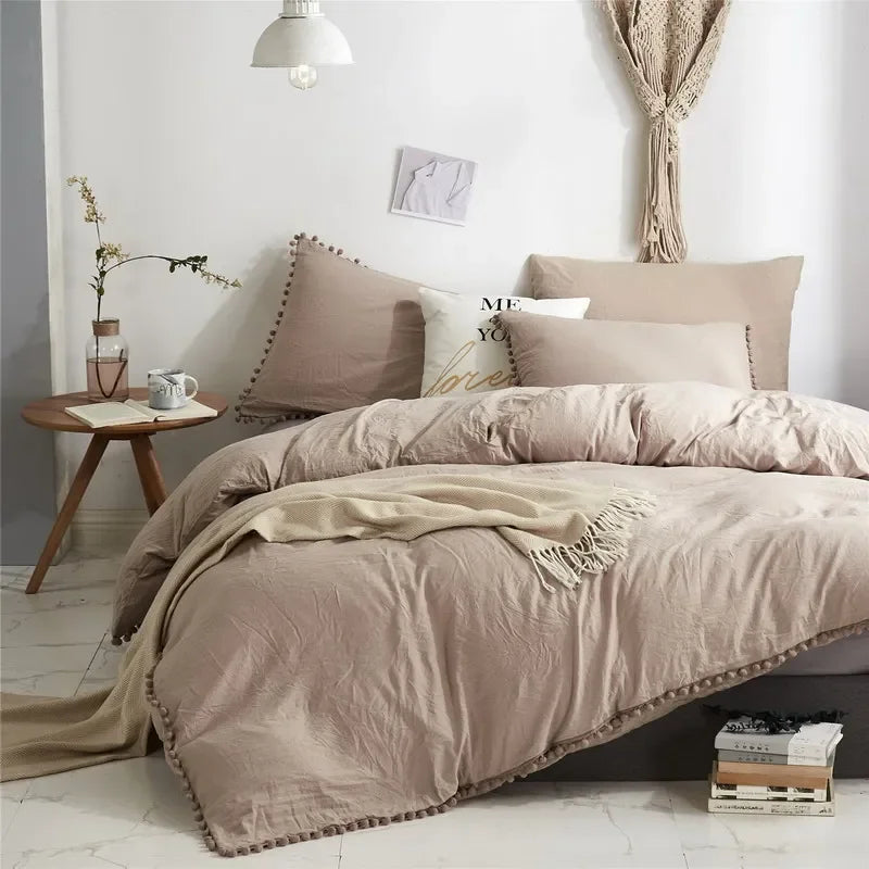 A beige duvet cover set with a pom-pom trim displayed on a bed in a bright room.