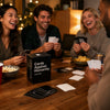 Four adults laughing while playing a card game on a wooden table with cards and game boxes.