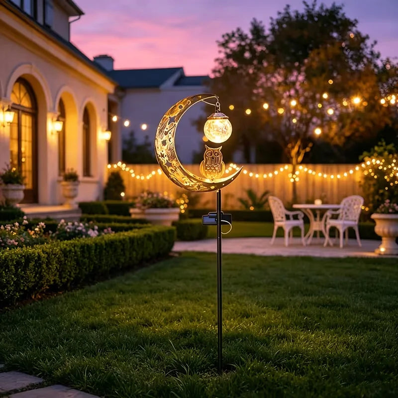 A solar owl garden light with moon decor hangs over a lawn in front of a house at dusk.
