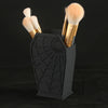 A black coffin-shaped makeup brush holder with a spider web design, holding three gold-handled makeup brushes.