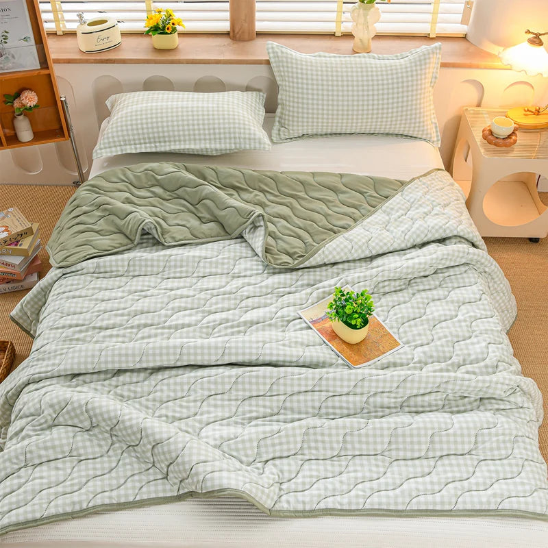 A light green quilted blanket with a white grid pattern is spread on a bed, next to a white pillow and wooden furniture.