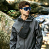 A man wearing a gray jacket and blue sunglasses stands outdoors near rocks, holding a black sling bag.