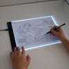 A hand draws on a transparent LED tracing light for artists, illuminating the lines on a white surface.