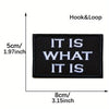 Black embroidered patch with white text IT IS WHAT IT IS on a fabric background