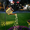 A vintage solar watering can garden light with a shepherd's hook illuminates a cascade of fairy lights over purple flowers in