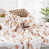 A white muslin baby swaddle blanket with a rainbow pattern is draped over a surface next to a woven basket.