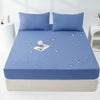 Una funda impermeable de colchón azul claro colocada en una cama con almohadas a juego y un vaso de agua.