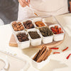 A clear plastic container with compartments holds various dried spices and herbs, being prepared by a hand.