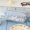 A light blue and white striped cosmetic pouch with a white bow and lace trim resting on a blue table.