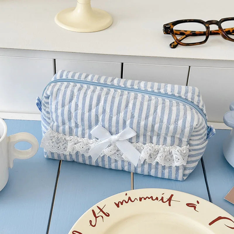 A light blue and white striped cosmetic pouch with a white bow and lace trim resting on a blue table.