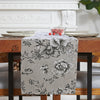 A gray floral patterned table runner displayed on a wooden table with white plates and decor in the background.