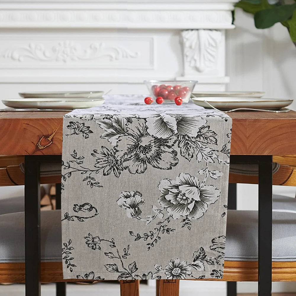 A gray floral patterned table runner displayed on a wooden table with white plates and decor in the background.