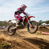 A motocross rider wearing pink and white gear jumps on a dirt bike in mid-air during an off-road race.