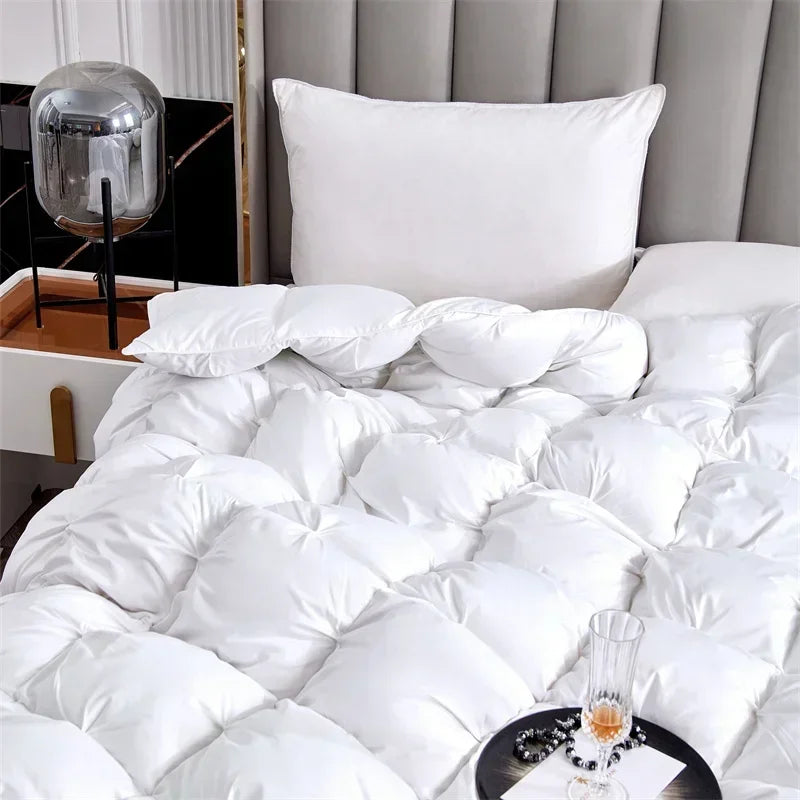 A white goose down comforter is shown with various pillows and a glass on a dark surface.