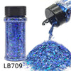 A bottle of blue holographic chunky nail glitter mix with a black shaker top and loose glitter spilling out.