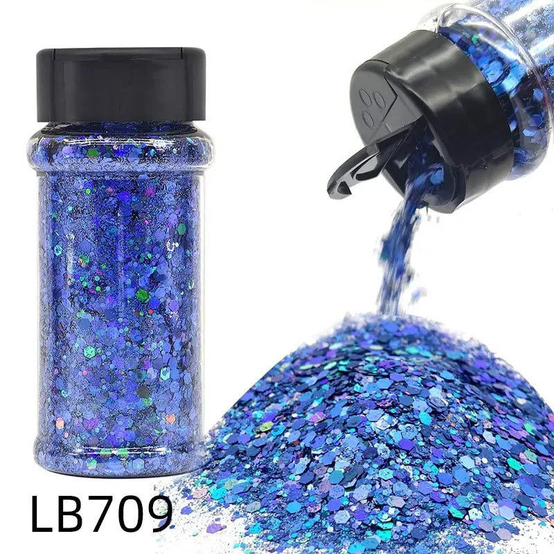 A bottle of blue holographic chunky nail glitter mix with a black shaker top and loose glitter spilling out.