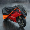 Une moto rouge est recouverte d'une housse de protection orange et noire tout en roulant sur une route mouillée.