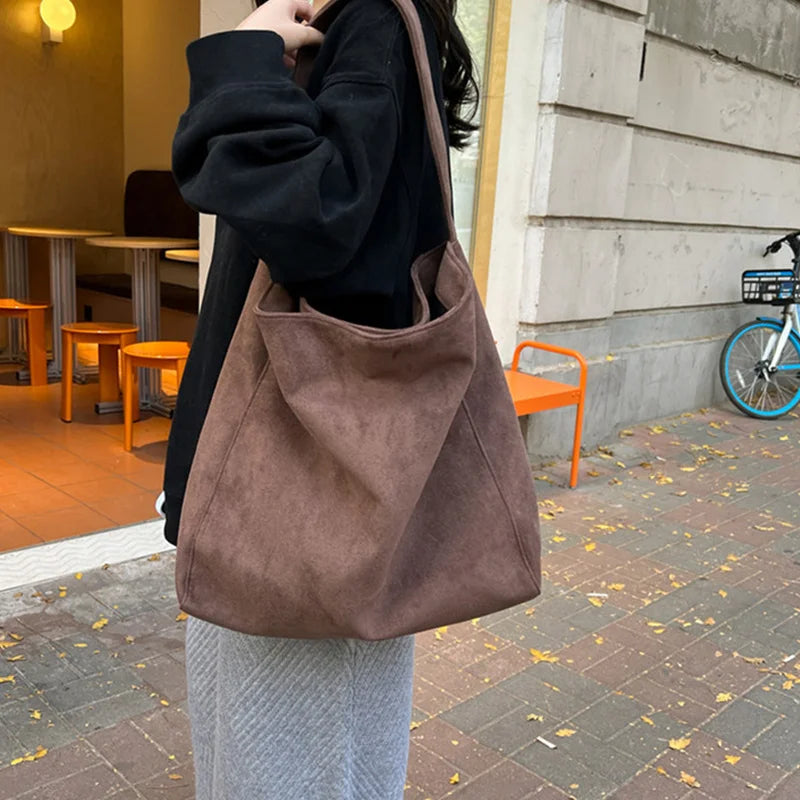 A person is holding a brown suede tote shoulder bag with a black sweater and light gray skirt outdoors.