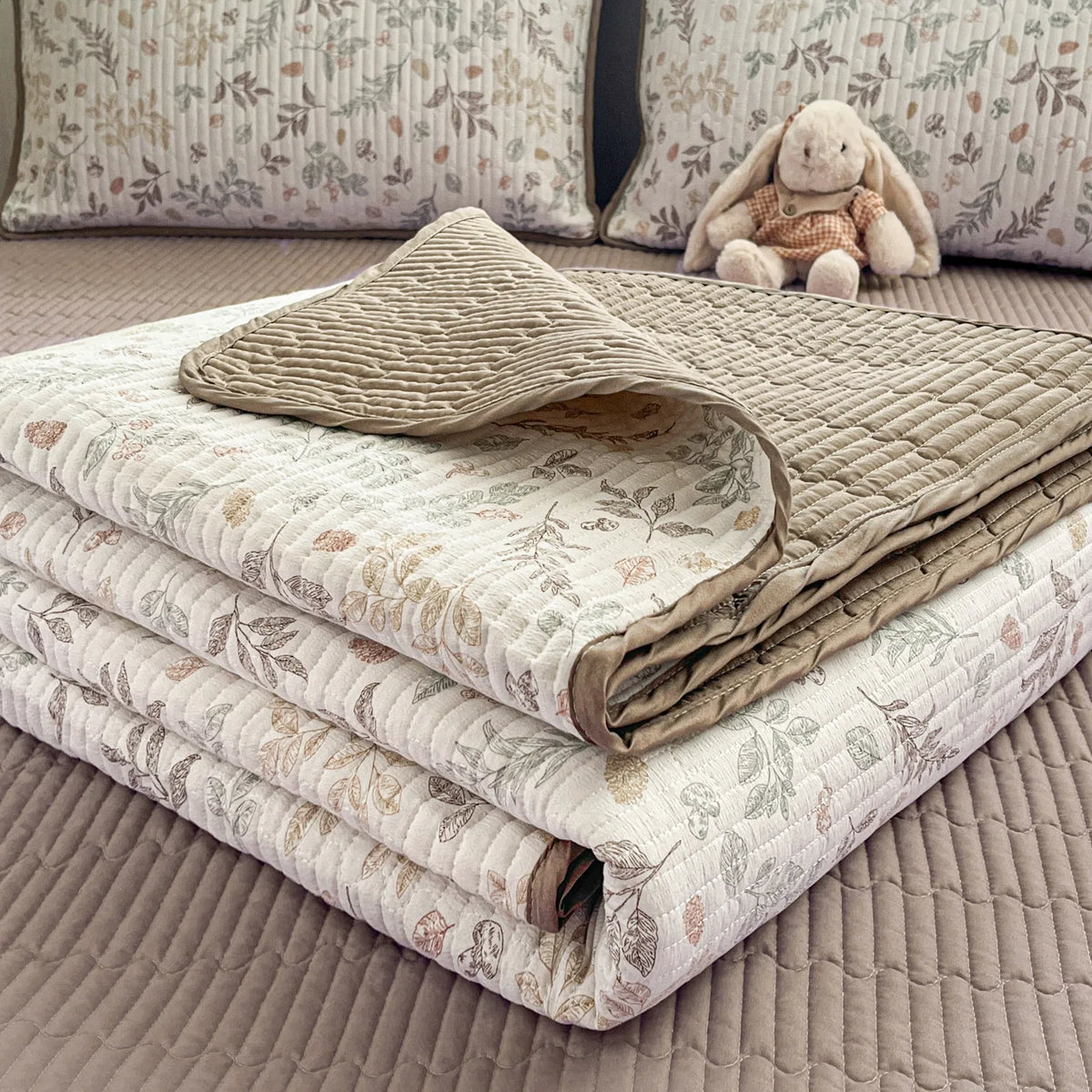 A stack of floral quilted bedspreads in beige and cream tones with a textured cover resting on a neutral surface.