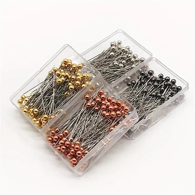 A clear plastic box contains a pack of precision quilting pins in various colors including gold, silver, bronze, and black.