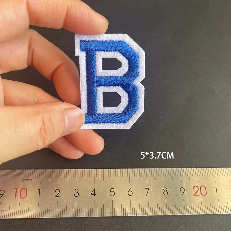 A hand holds a blue embroidered letter B patch, measuring 5x3.7 cm against a ruler.