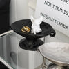A black ceramic soap dish with an elevated stand holds a white dove figurine and gold rings.