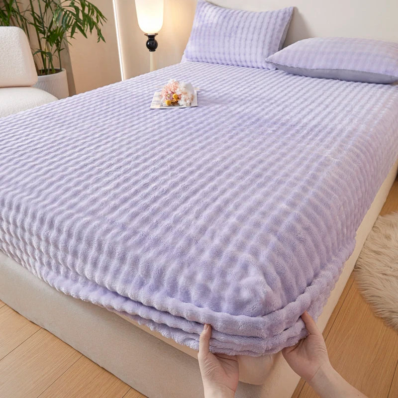 A purple faux fur fitted sheet is draped over a bed, with a hand holding the bottom edge.