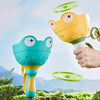 A hand is holding a yellow and green pull-string flying saucer launcher toy for kids against a scenic outdoor background.