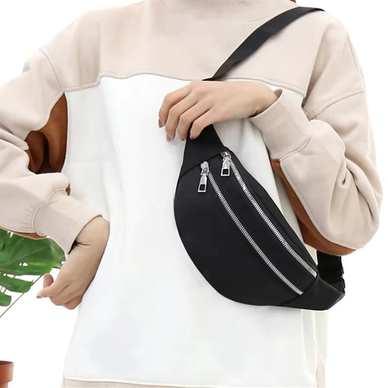 A close-up of a black nylon waist pack with a zipper and adjustable strap worn on a light beige sweater.