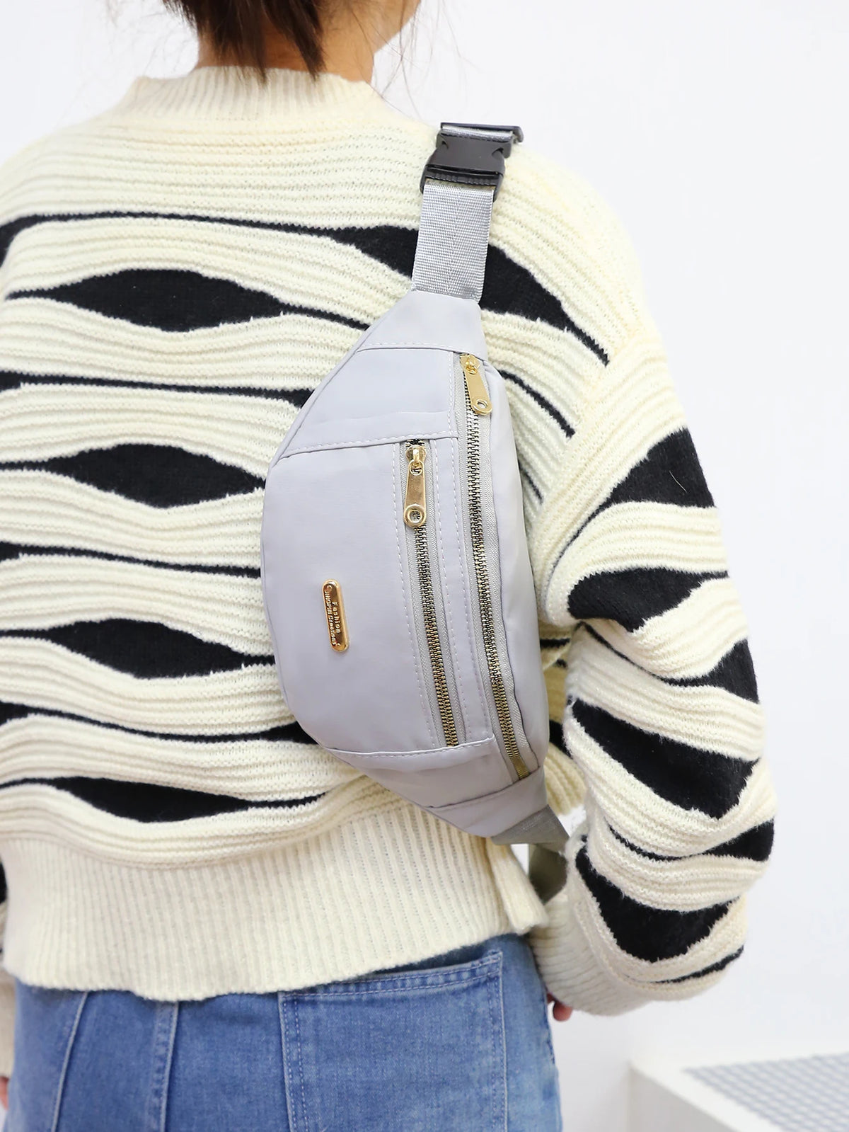A person wearing a cream striped sweater is shown from the back, carrying a light gray nylon waist pack on their shoulder.