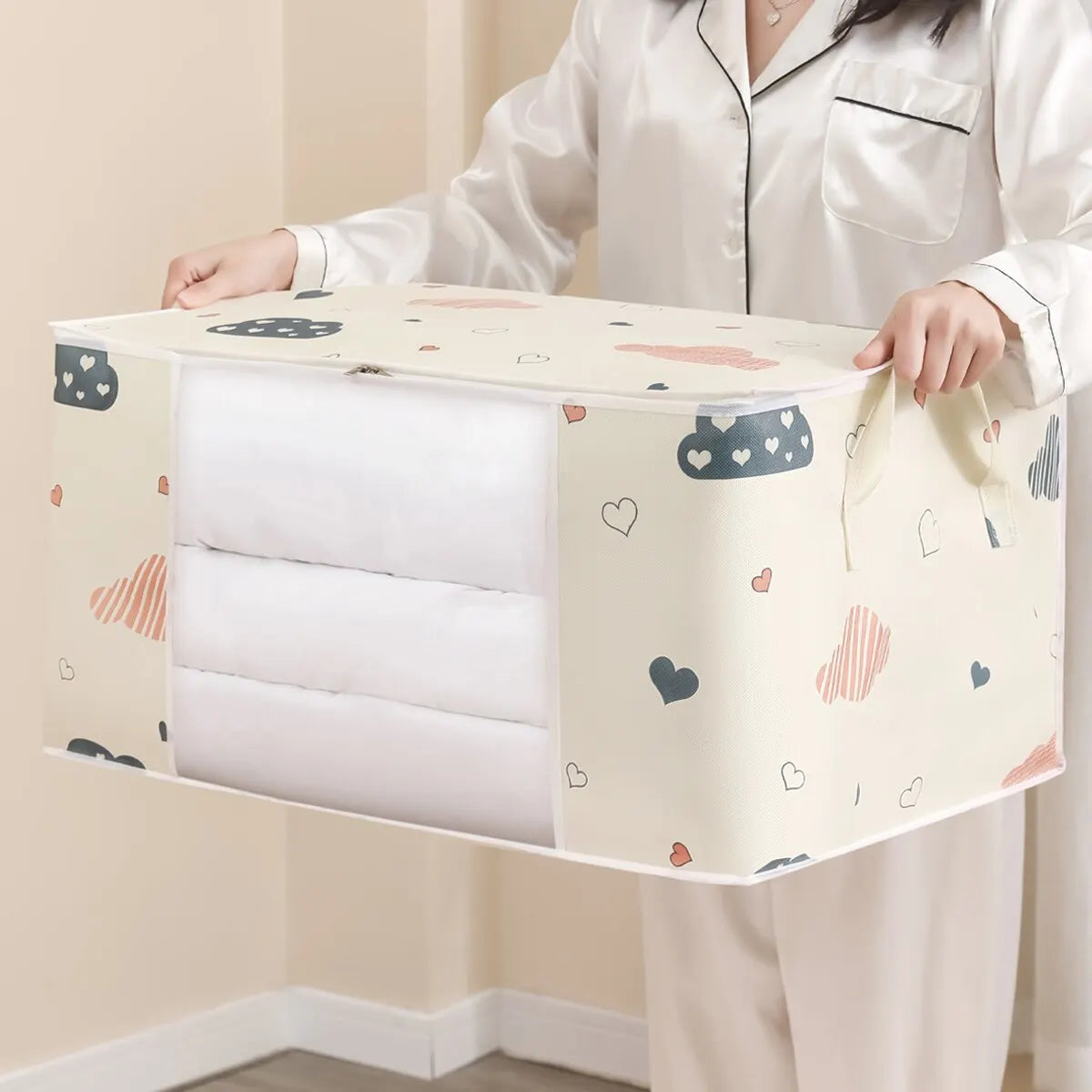 A person holds a beige foldable blanket and clothes storage bag with a zipper, featuring a light background and small heart a