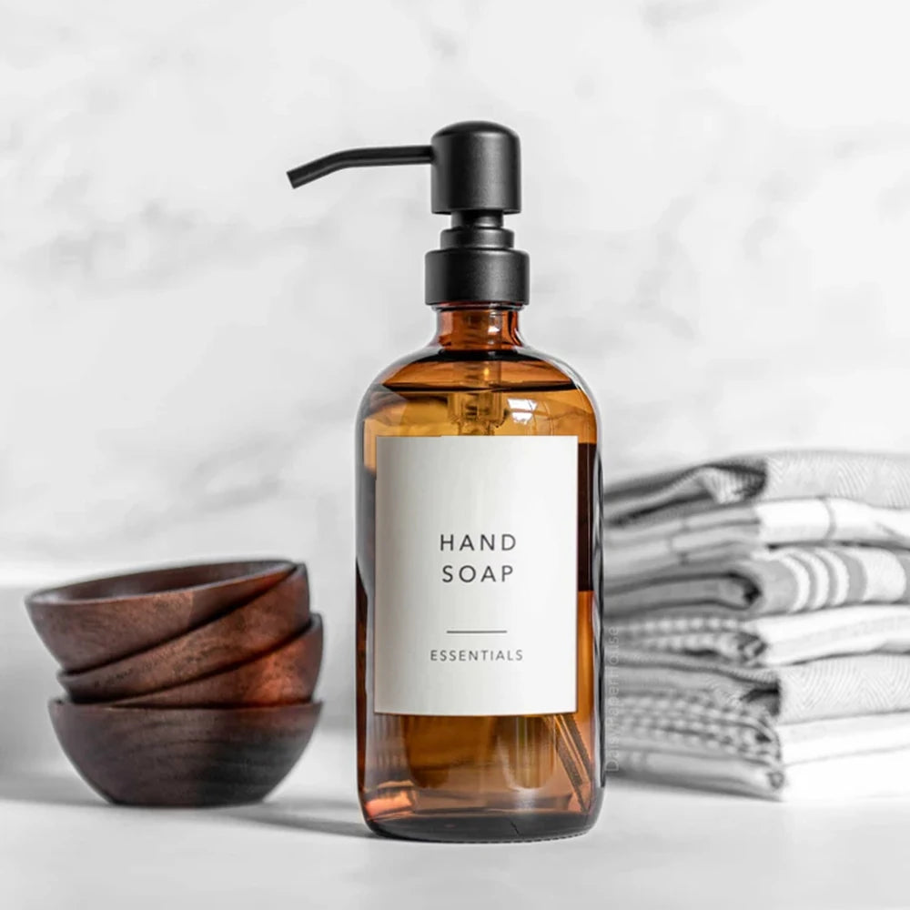 Amber glass hand soap dispenser bottle with a black pump, set against a white marble background and stacked towels.