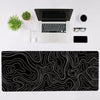 A black topographic map design mouse pad with white lines, a silver laptop, and accessories on a white desk.