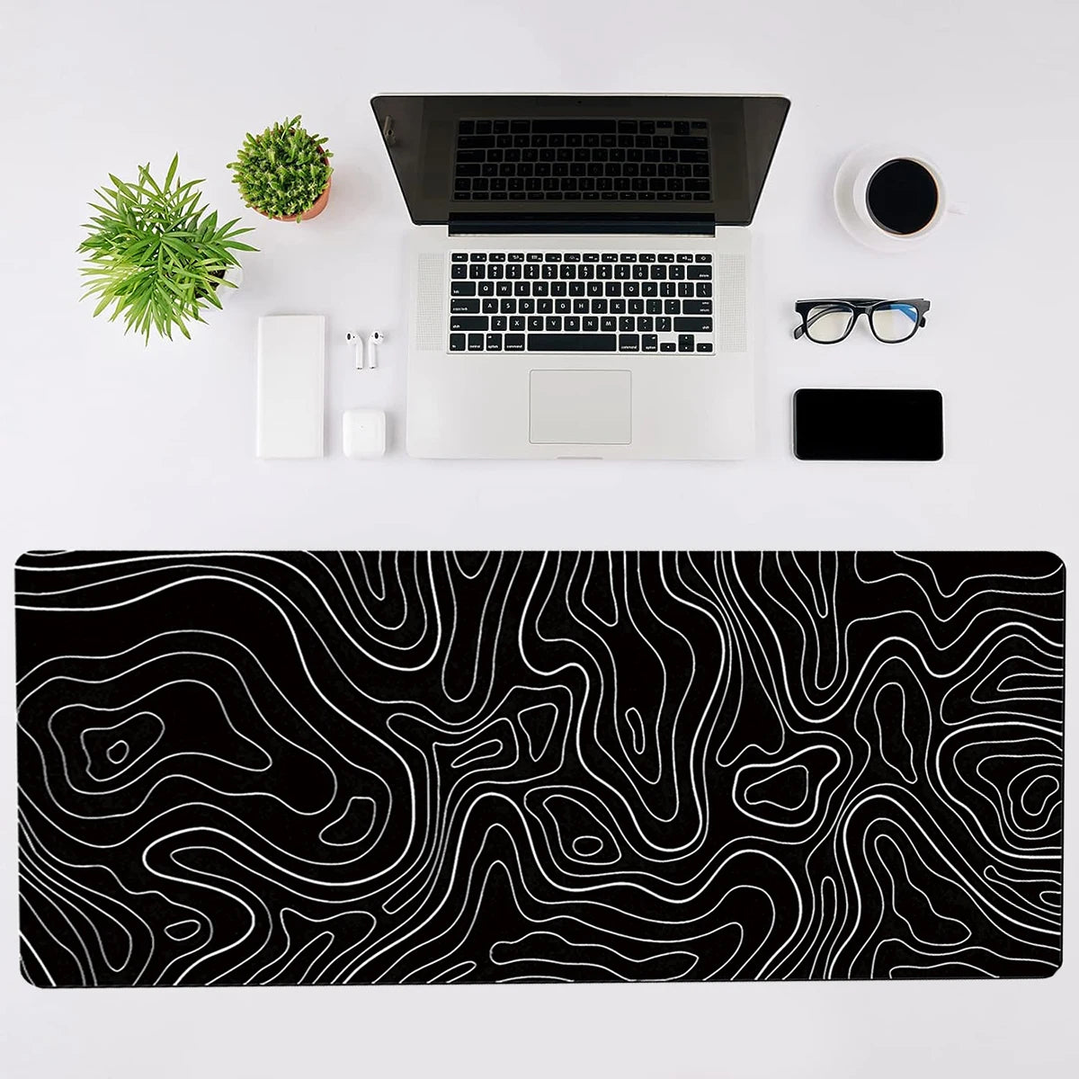A black topographic map design mouse pad with white lines, a silver laptop, and accessories on a white desk.