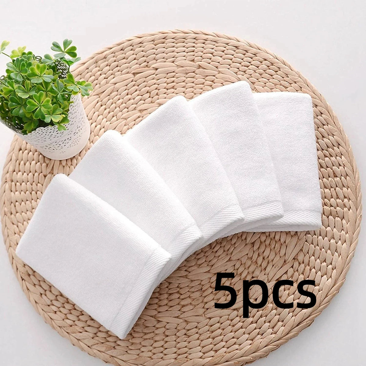 Five white soft cotton terry cloth face towels arranged on a woven mat next to a small green plant.