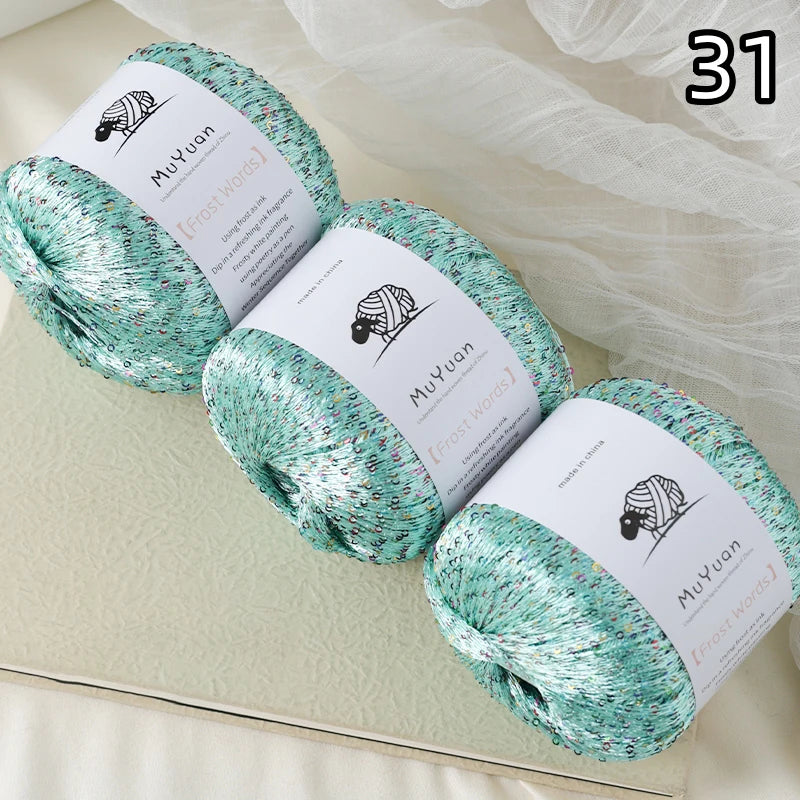 Three skeins of turquoise glitter yarn labeled MuYuan Frost Words displayed on a light surface.