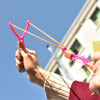 A hand holds a pink LED glowing flying arrow toy with rubber band launchers against a blue sky.
