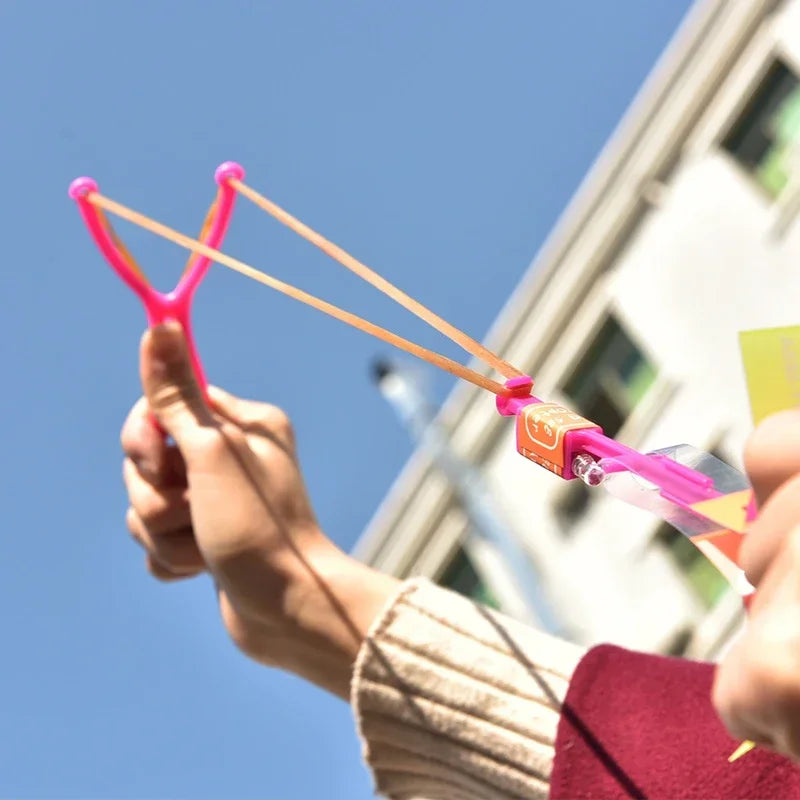A hand holds a pink LED glowing flying arrow toy with rubber band launchers against a blue sky.