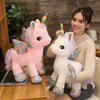 A young woman is holding a white unicorn plush toy with rainbow accents, set against a wooden background.