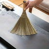 A hand is holding a natural grass broom with a woven handle, positioned over a gray wooden table.