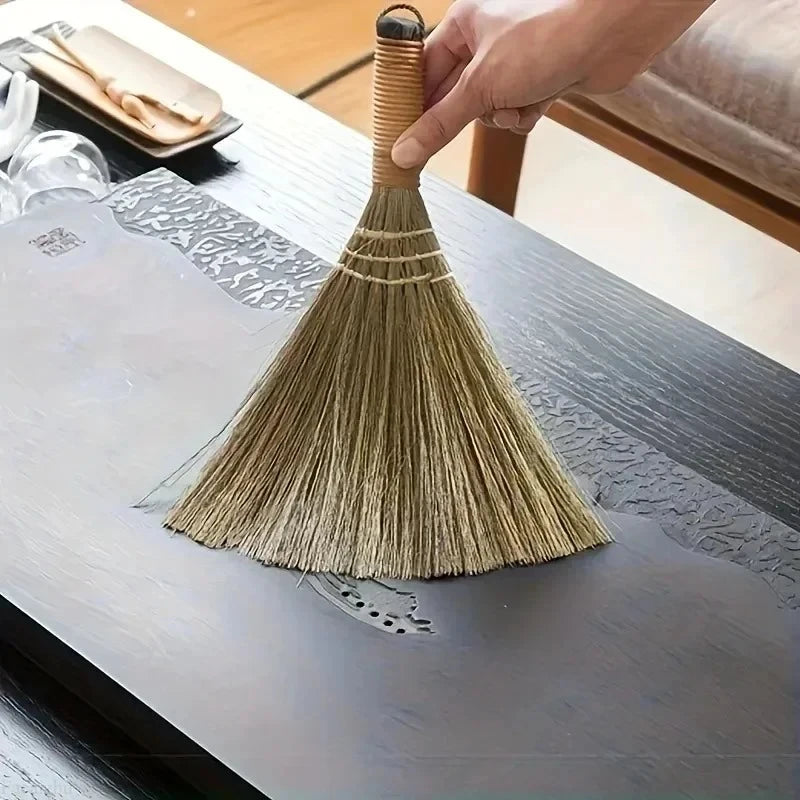A hand is holding a natural grass broom with a woven handle, positioned over a gray wooden table.
