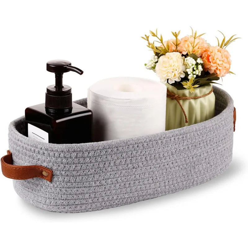 A woven cotton rope storage basket holding a black soap dispenser, a white roll of paper towels, and a bouquet of orange and 