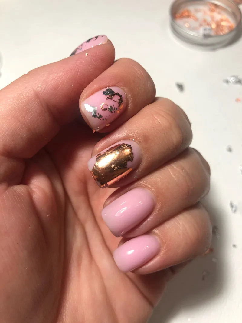A hand displays a manicure featuring pink and gold metallic foil flakes on the nails.