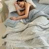 A woman rests on a bed covered with a striped light blue quilt and pillows.