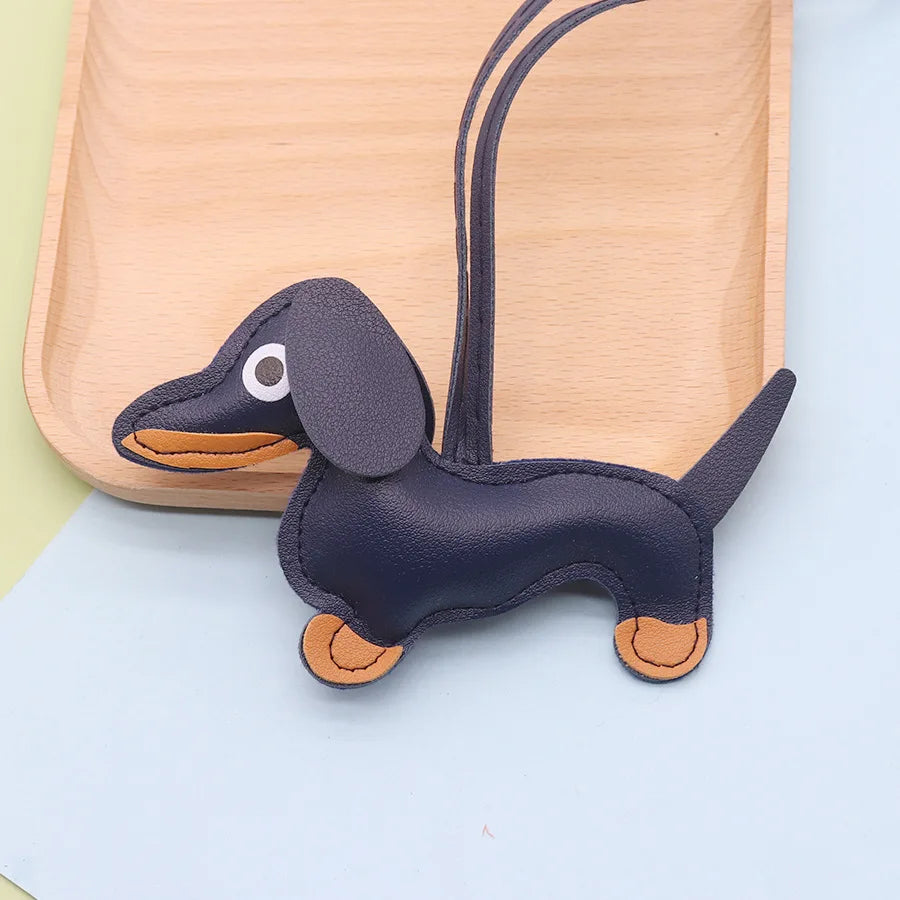 A dark gray dachshund shaped PU leather keychain with an elongated body and floppy ears resting on a light wooden board.